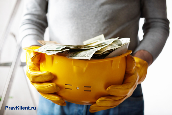 Рабочий держит в руках строительную каску с деньгами